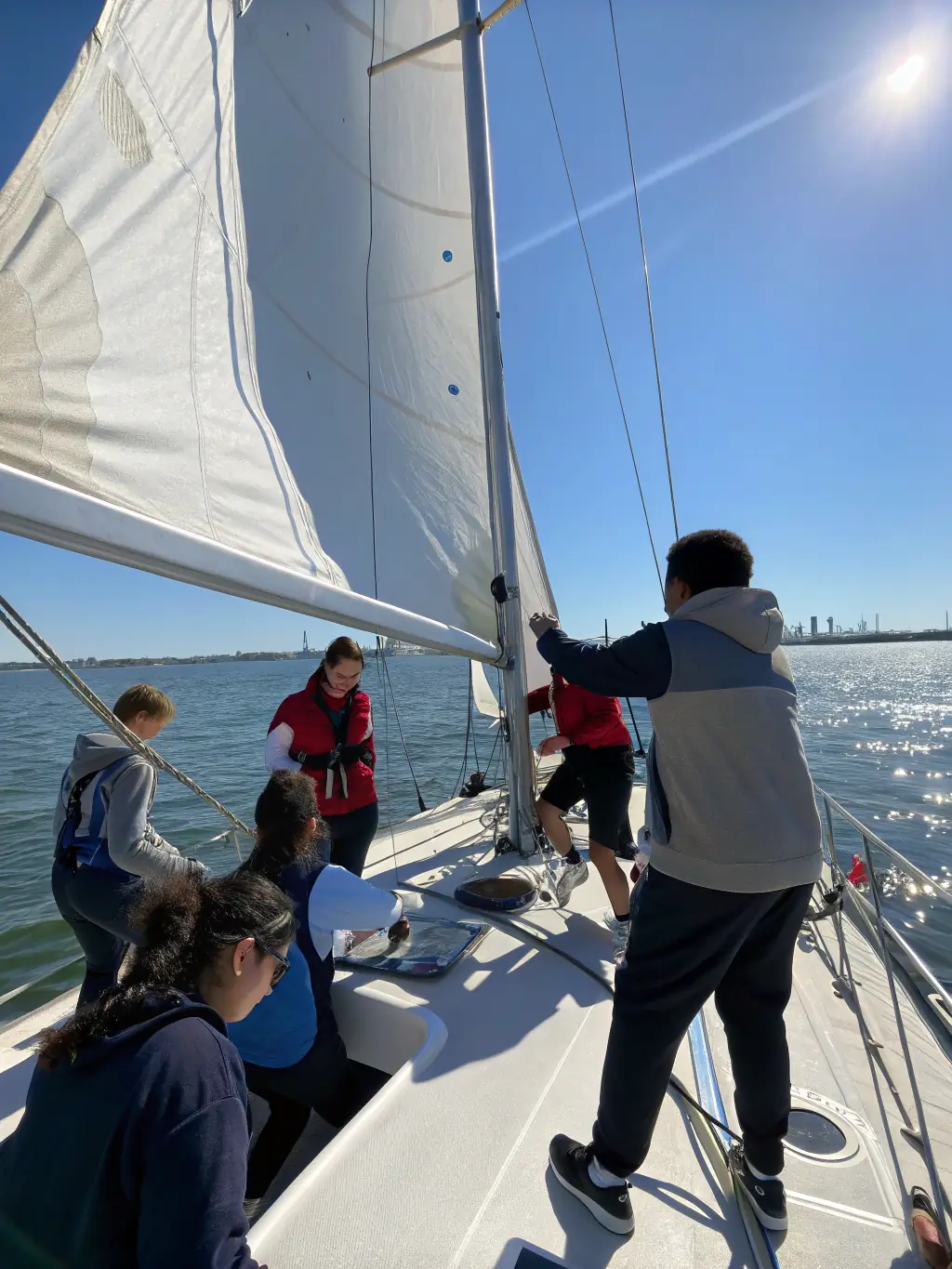 A group of diverse sailors laughing and working together to raise the sail on a sailboat during a sunny day at CLUB NAUTIQUE SAINT AMANDOIS - BASE DE VIRLAY, emphasizing teamwork and camaraderie.